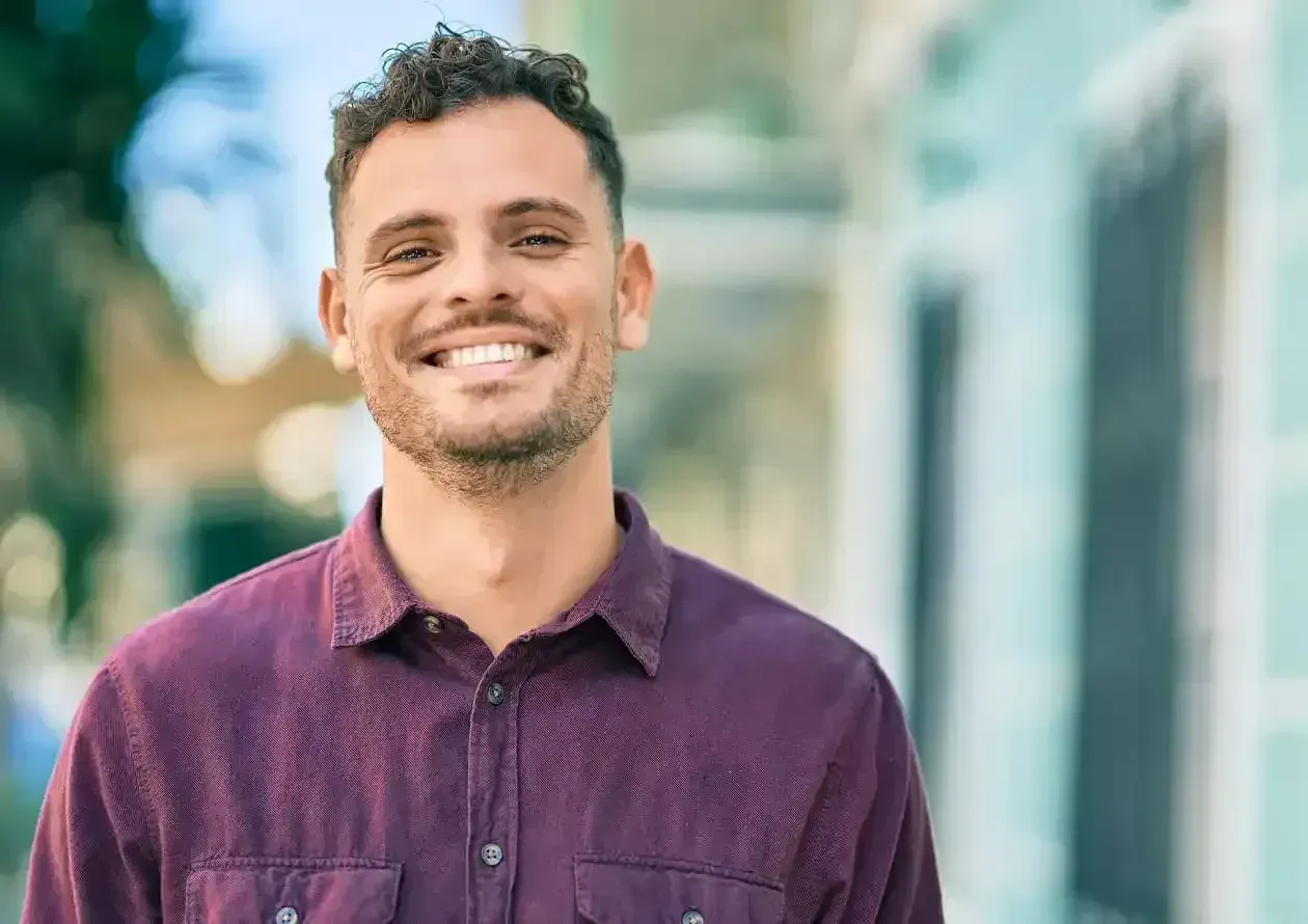 Smiling man with curly hair and beard wearing a maroon button-up shirt outdoors.